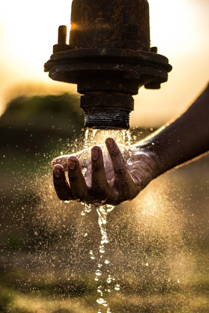 pexels-photo-2837863 A person washes their hand under a rustic water pipe, capturing the essence of fresh water at sunset.